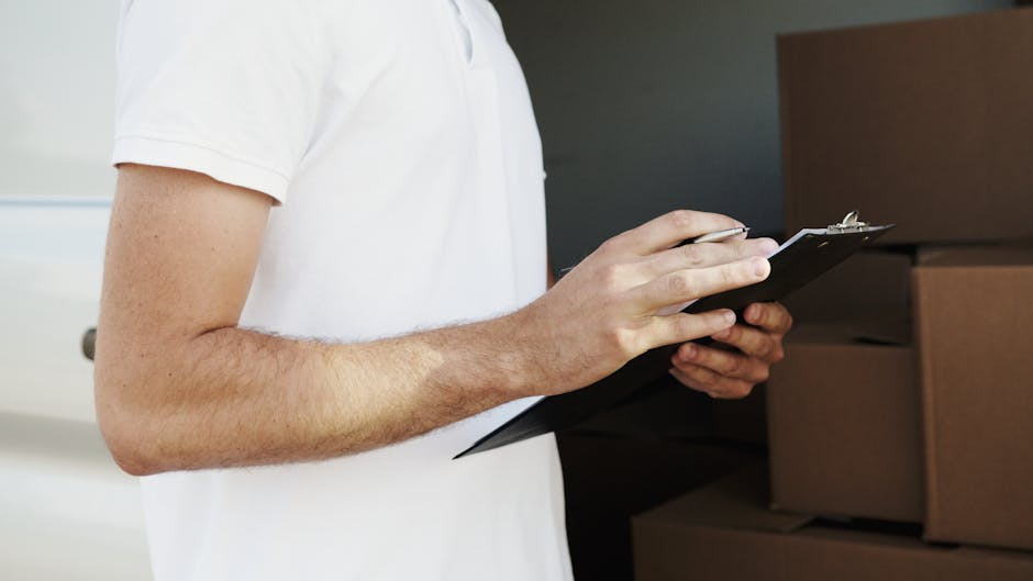 Close-up of a person wearing a white t-shirt holding a black clipboard and pen, standing inside a property near stacked cardboard moving boxes and brown packing crates. The individual is reviewing a moving inventory or checklist as part of a home relocation process, with the boxes arranged on the floor and a doorway visible in the background. The scene reflects furniture transport, packing, and loading activities associated with a professional removals service, such as those offered by Kensington Removals. The environment is well-lit, highlighting the details of the packing materials and the careful planning involved in a household move.