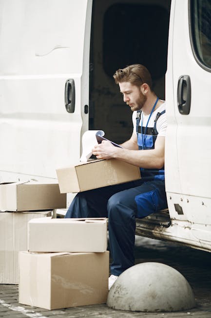 A male mover with short brown hair and a beard, wearing a blue and white striped shirt and blue work trousers, seated on the edge of a white van with the sliding door open. He is holding a clipboard and a pen while inspecting or recording details. Around him on the pavement are several cardboard boxes, some of which are open and partially packed, with brown packing paper visible inside. The scene depicts a home relocation process, with the man preparing or checking items for transportation, and the van positioned near a building entrance, suggesting a residential moving service possibly provided by Kensington Removals. The environment is well-lit, with natural daylight, and the background shows a clean, urban setting with a rounded concrete bollard nearby.