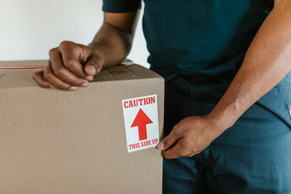 Close-up of a person wearing a white t-shirt holding a black clipboard and pen, standing inside a property near stacked cardboard moving boxes and brown packing crates. The individual is reviewing a moving inventory or checklist as part of a home relocation process, with the boxes arranged on the floor and a doorway visible in the background. The scene reflects furniture transport, packing, and loading activities associated with a professional removals service, such as those offered by Kensington Removals. The environment is well-lit, highlighting the details of the packing materials and the careful planning involved in a household move.