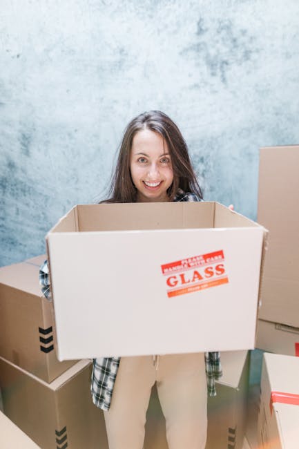 A man and a woman standing at the entrance of a property after a home relocation, both holding cardboard boxes used for packing. The man, dressed in a maroon t-shirt and orange pants, appears to be lo