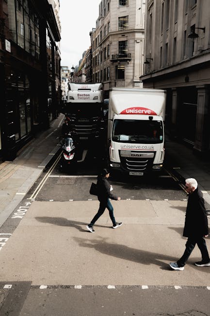 An urban street scene showing a narrow road with two delivery trucks parked perpendicular to the curb, one branded with uniserve.co.uk and the other with Becton Transport. A small motorcycle is parked
