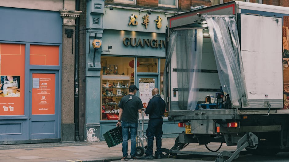 An urban street scene showing a narrow road with two delivery trucks parked perpendicular to the curb, one branded with uniserve.co.uk and the other with Becton Transport. A small motorcycle is parked