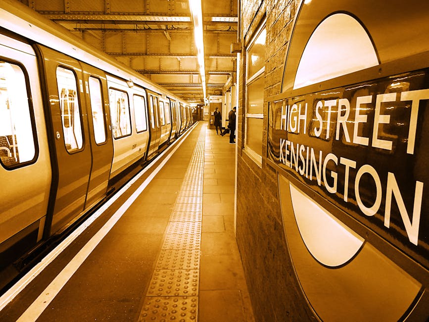 Inside Kensington Olympia underground station platform with a train arriving or departing on the left side. The platform is illuminated with warm lighting and has a tactile yellow safety strip along the edge. To the right, a station sign displays 'High Street Kensington' in bold white letters on a black background, mounted on a wall with a large circular design panel. In the background, a few people are near the platform, some walking and others waiting, with luggage or bags visible, indicating a setting related to home relocation or moving logistics. The scene portrays a typical scene of travel and transit, relevant to house removals and furniture transport services offered by Kensington Removals, emphasizing organized, professional logistical handling of transportation and packing during moves.
