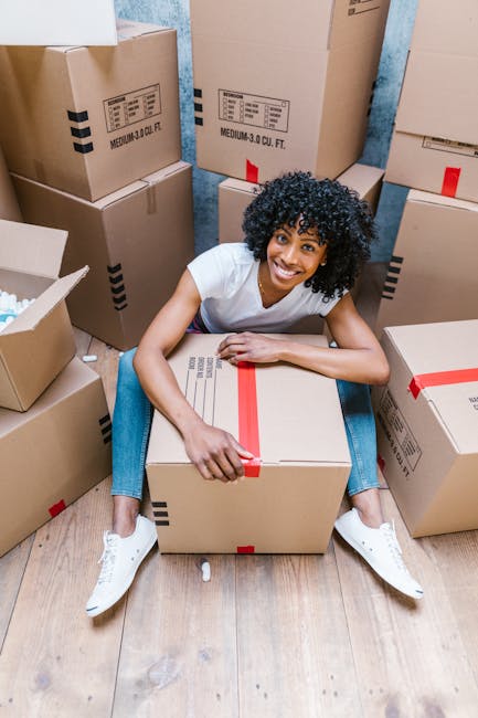 A woman with curly black hair, wearing a white T-shirt and blue jeans, sits on a wooden floor surrounded by numerous cardboard boxes of varying sizes, some sealed with red packing tape. She is smiling and holding a large cardboard box in her lap. The boxes display printed labels and are positioned both on the floor and stacked against a wall covered with blue wallpaper. In the background, there are additional boxes and packing supplies, indicating an active packing and moving process. The scene reflects the preparation phase of home relocation or furniture transport, with a focus on packing and organizing household items for a move, and illustrates professional removals service equipment such as boxes and packing materials.
