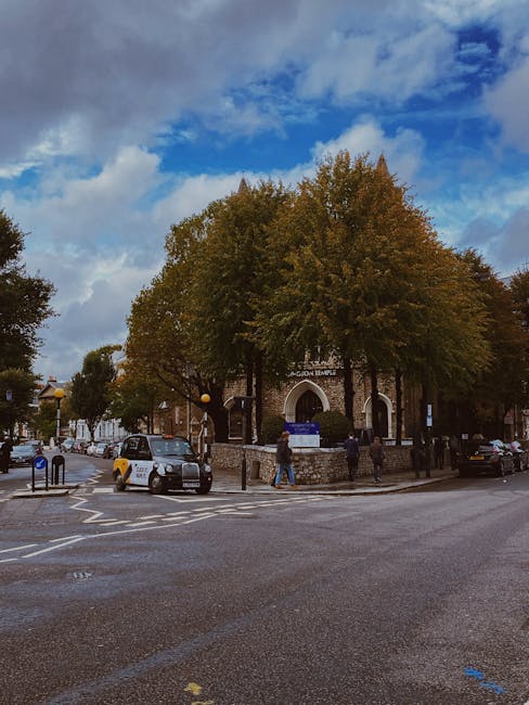 Photograph of a street scene outside a church building with the sign 'Kensington Removals' visible on a blue board in front of the entrance. The church is partially obscured by large, leafy trees with green and brown foliage, indicating autumn. Several cars are parked on the street and pavement, including a silver vehicle near the curb, and there are road markings and bollards visible in the foreground. People are walking across the street and along the sidewalk outside the church, some engaged in lifting or carrying boxes wrapped in plastic or cardboard, suggesting activities related to house removals or furniture transport as part of a home relocation process. The sky is partly cloudy with patches of blue, and streetlights can be seen along the road. The scene captures a typical urban environment during daylight hours, emphasizing moving logistics near a community building, with Kensington Removals providing professional relocation support.