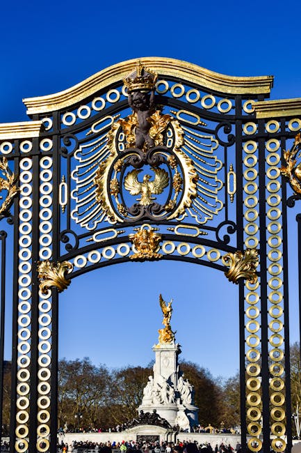 A detailed view of an ornate, black and gold decorative gate with circular and floral motifs, topped by a sculpted figure of a lion. Behind the gate, a bright blue sky is visible along with a distant statue on a tall pedestal featuring a winged figure. The scene appears to be outdoors, potentially at a historic or cultural site, with trees visible in the background. This image captures the elaborate craftsmanship and grandeur of the entrance, which could be part of a public park or heritage landmark, often associated with house removals and relocation services such as those offered by Kensington Removals when moving into or out of heritage properties or historic districts.