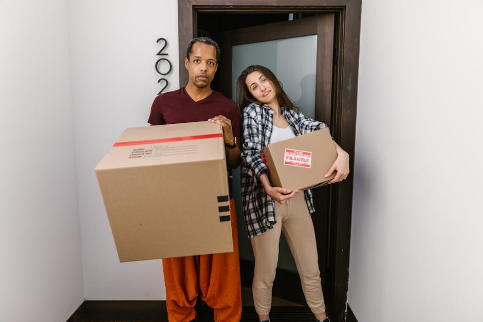 A man and a woman stand in a doorway inside a home, each holding a cardboard moving box. The man, dressed in a maroon t-shirt and orange trousers, has a serious expression and is holding a large box with red and black tape, indicating it is part of packing and moving activities. Behind them, the woman, wearing a plaid shirt over a white top and beige trousers, leans her head slightly to the side with a relaxed expression while holding a smaller box marked with a 'Fragile' label. The doorway features a dark wooden frame, and the interior walls are white, highlighting a typical home environment prepared for a house removal. The scene reflects the process of furniture transport and packing during a home relocation, with visible packaging materials and the careful handling of contents as part of a professional removals service, such as that provided by Kensington Removals.