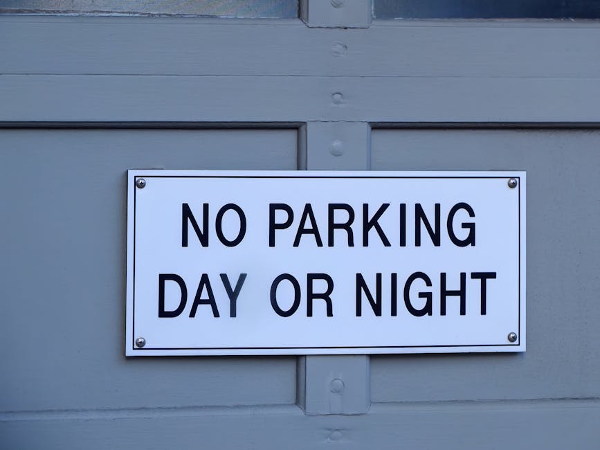 A rectangular white sign with bold black text reading 'NO PARKING DAY OR NIGHT' is mounted on a grey wooden door or wall. The sign is secured with four silver screws, one in each corner. The background surface has visible horizontal and vertical paneling, typical of an exterior dwelling or property entrance. The lighting is neutral, with no shadows or reflections, indicating natural or soft indoor light. This sign, displayed outside a property, is related to parking regulations in relation to house removals or moving services, such as those offered by Kensington Removals, highlighting restrictions during logistical moving operations and adherence to local permit rules for furniture transport or home relocation within the Kensington area.