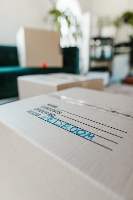 A close-up of a cardboard moving box labeled 'BEDROOM' in blue text, situated on a surface in a well-lit living room. The background shows other packed boxes, a sofa, a coffee table, and some househol