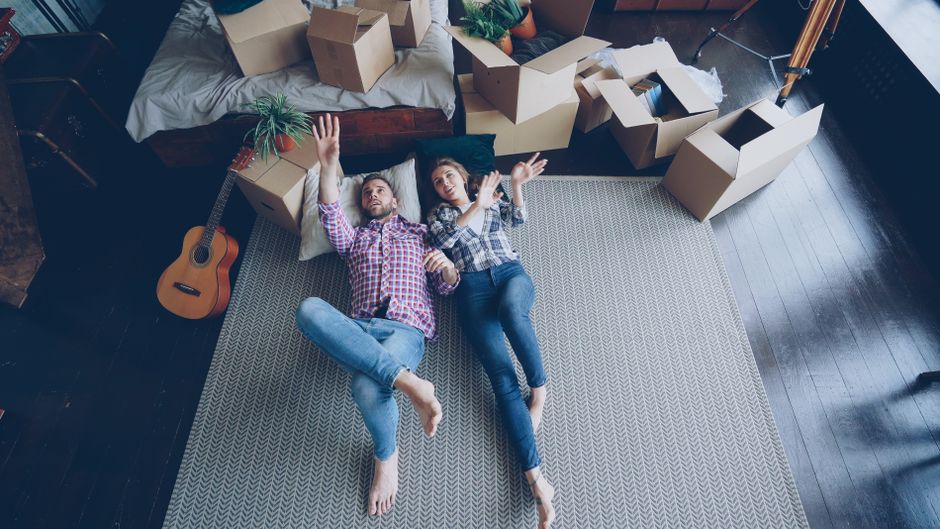 A close-up of a cardboard moving box labeled 'BEDROOM' in blue text, situated on a surface in a well-lit living room. The background shows other packed boxes, a sofa, a coffee table, and some househol