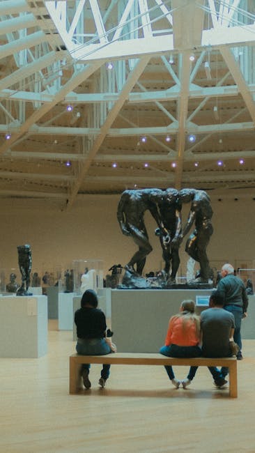 Inside an art gallery or museum with a curved wooden ceiling structure and large skylights, several bronze sculptures of human figures are displayed on platforms. The sculptures depict two figures bent over, appearing to be engaged in a task or interaction, with other smaller sculptures visible in the background. In the foreground, three visitors—two children and an adult—are seated on a wooden bench facing the sculptures, with their backs to the camera. The scene includes a smooth, polished wooden floor, and the lighting appears to be natural, supplemented by ceiling lights. This setting likely relates to art curation or exhibition preparation, and the sculpture placement may be part of a professional moving or handling process involving careful packing and transport, supported by Kensington Removals' specialization in house and art relocation, particularly for museums and exhibitions.